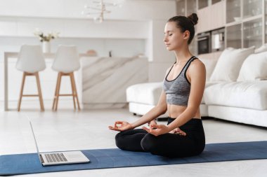 Oturma odasındaki egzersiz minderinde meditasyon yapan, online eğitim talimatlarını tekrarlayan, video konferans yoluyla sanal yoga dersi için laptop kullanan sağlıklı genç bir kadın. Zindelik