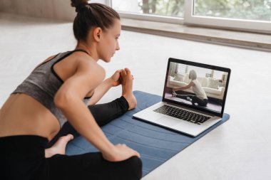 Spor giyim bölümünde dizüstü bilgisayar kullanarak yoga yapan formda bir kadın. Bayan koçun bilgisayarında sanal yoga dersi var. Sağlıklı ve aktif yaşam tarzı konsepti. Çevrimiçi ev eğitimi