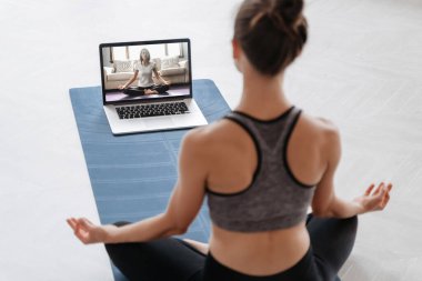 Evde dizüstü bilgisayarla yoga yapan genç ve sportif bir kadının yakın çekimi. Yoga eğitmeni video konferansı aracılığıyla video eğitimi veriyor. Meditasyon, Sukhasana, rahatla. Sağlıklı yaşam tarzı kavramı