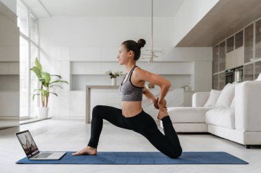 Toplumsal uzaklık nedeniyle evde antrenman yapan, minder üzerinde yoga yapan sağlıklı, genç bir kadının tam boy çalışması sanal egzersiz sınıfına sahip, esneme. Çevrimiçi fitness, sağlık ve sağlık konsepti