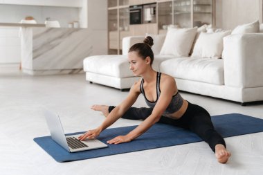 Oturma odasında paspas üzerinde gerinen, sanal eğitim için dizüstü bilgisayar kullanan, egzersiz tekrarlayan formda genç bir kadının çevrimiçi fitness dersi var. Sağlık, sağlıklı yaşam tarzı konsepti 