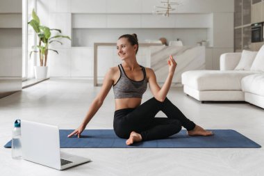 Kadınlar sağlık ve sağlıklı bir vücut için oturarak büküm egzersizleri yapıyorlar. Evde yoga, fitness, egzersiz konsepti. Dizüstü bilgisayarda ders izleyen gülümseyen güzel bir kızın evinde yoga dersi var.