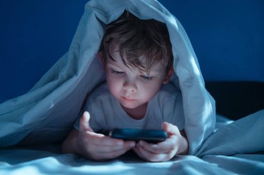 Küçük çocuk gece battaniyenin altında yatar, akıllı telefon tutar, mobil oyunlar oynar, sosyal medyayı kaydırır ya da çizgi film izlerdi. Çocuklar arasında dijital bağımlılık ve aşırı ekran zamanı alışkanlıkları. 