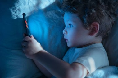 Gece yatağında uzanmış, akıllı telefona dikkatle bakan mavi ekran ışığıyla aydınlatılmış içeriğe dalmış küçük bir çocuğun üst görüntüsü. Gadget bağımlılığı ve uzun ekran aşırı kullanımı.