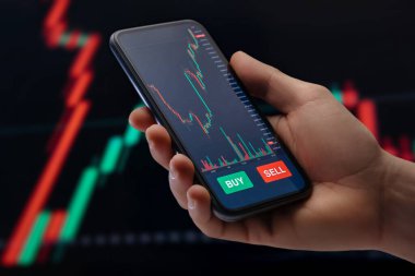 Kapat, akıllı telefonu elinde tutan tüccar ekranda bir mobil ticaret platformu uygulaması, şamdan şeması ve arka planda sermaye veya kripto yatırımları için finansal grafikler.