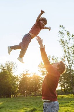 Baba neşeli oğlunu bir parkta havaya fırlatıyor, gülen çocuk güneşli bir yaz gününde babasıyla dışarıda eğleniyor. Aile eğlencesi, bağlanma ve Babalar Günü konsepti. Boşluğu kopyala