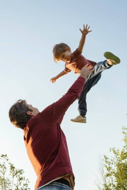 Baba ve oğul parkta birlikte oynuyorlar, baba gülücük saçan oğlunu havaya fırlatıyor, eğleniyor ve ailece vakit geçiriyorlar. Mutlu çocukluklar ya da Babalar Günü konsepti. Metin için boşluk