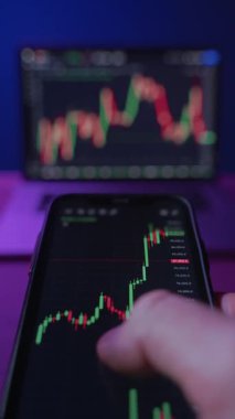 Akıllı telefon tutan tüccara yakın plan, ticari uygulama kullanarak kripto ve borsa trendlerini analiz ederek neon ışıklı ofisinde arka planda birden fazla monitörle. Dikey video