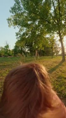 Güneşli bir günde gökyüzü ve ağaçlarla kaplı bir ebeveyn tarafından terk edilirken neşeli bir şekilde gülümseyen heyecanlı genç çocuk. POV bakış açısı, bir babanın yüksekten atması ve gülen oğlunu parkta yakalaması. Dikey video