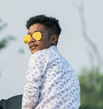 Arkadan kameraya bakan genç bir adam altın güneş gözlüğü takıyor.