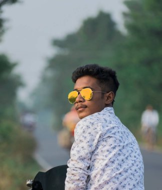 Arkadan kameraya bakan genç bir adam altın güneş gözlüğü takıyor.