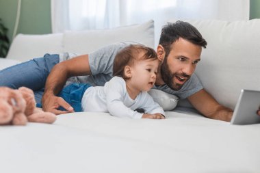 Şirin bebek oğlu tutan ve tablet vasıl bakarak mutlu gülümseyen beyaz adam.