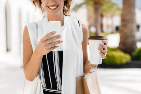 Cropped smiling Caucasian woman shopper typing on her smartphone and holding cup of coffee outdoors.