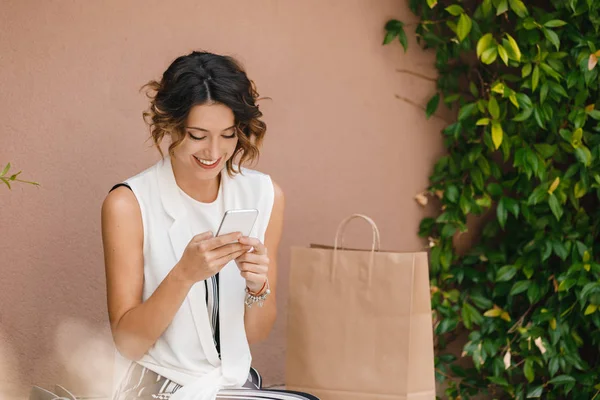 美しい笑顔の白人女性ショッピング バッグと屋外に座って 彼女の携帯電話で入力 — ストック写真