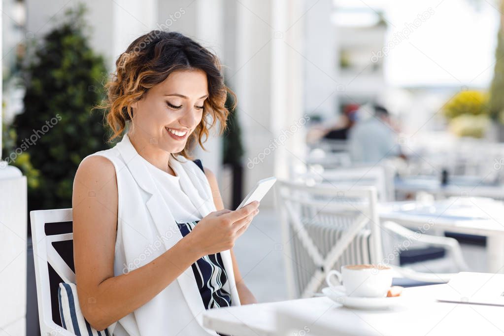 Pretty Caucasian smiling woman sitting at cafe and texting on her mobile phone.