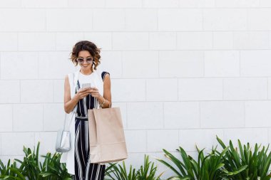 Güzel beyaz kadın holding alışveriş çantası gülümseyen ve cep telefonunu yazarak.