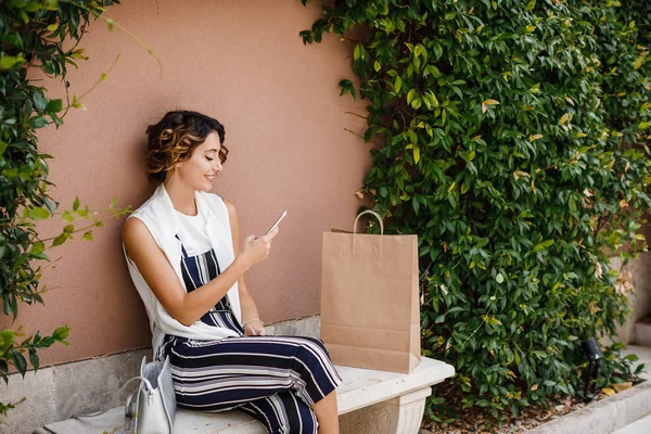 Beautiful smiling Caucasian woman shopper looking at her cell phone and sitting on bench.