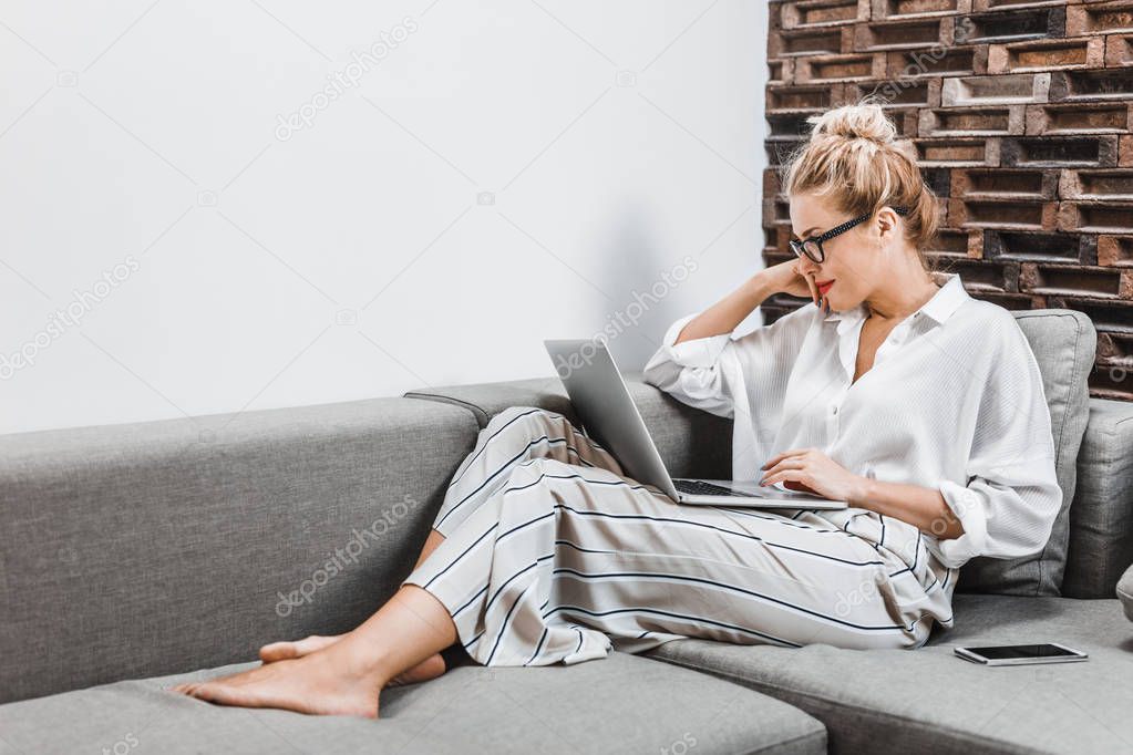 Beautiful blonde Caucasian woman sitting on couch at home and looking at laptop.