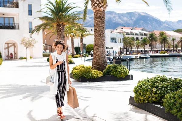 Cep telefonunu yazarak ve alışveriş çantası holding deniz tarafından yürüyüş güzel şık gülümseyen kadın.