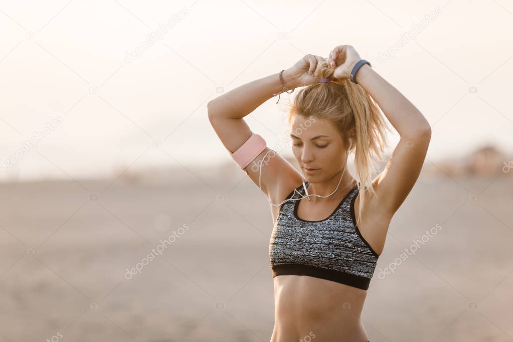 Pretty Caucasian sportswoman standing on sandy beach and holding her ponytail.