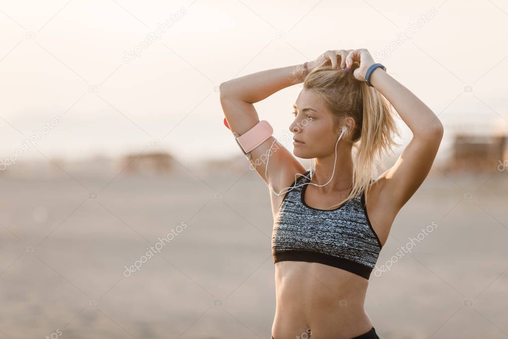 Pretty Caucasian sportswoman standing on sandy beach and holding her ponytail.