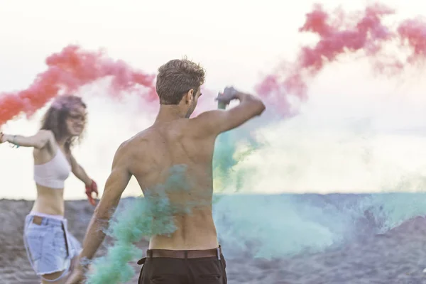 Young man and woman dancing with smoke bombs on beach.