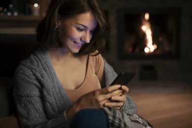 Şöminenin yanında oturan ve Smartphone'da yazarak gülümseyen güzel kadın.