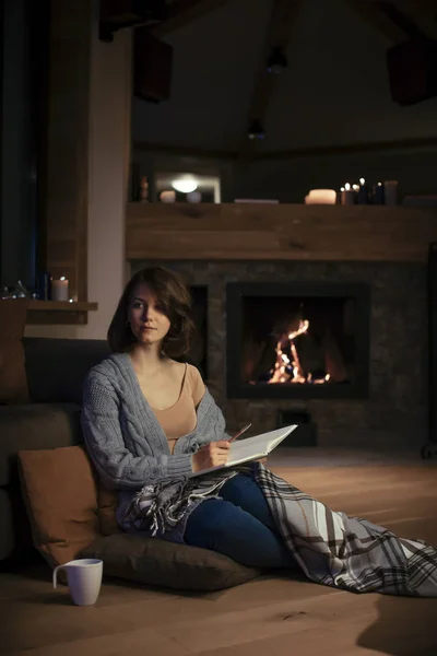 Beautiful woman sitting by the fireplace in cozy sweater and writing in notebook.