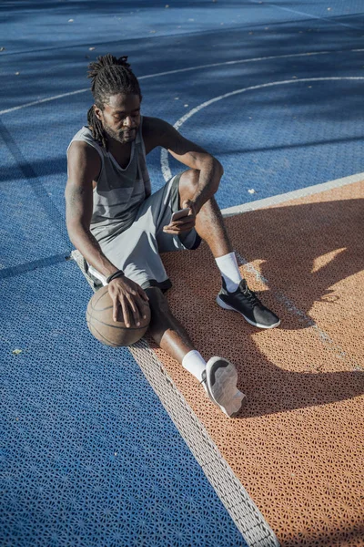 英俊年轻的黑人篮球运动员使用他的手机和举行篮球 而坐在一个户外篮球场 — 图库照片