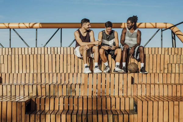 三英俊的朋友和篮球运动员在使用手机和坐在户外篮球看台上玩得很开心 — 图库照片