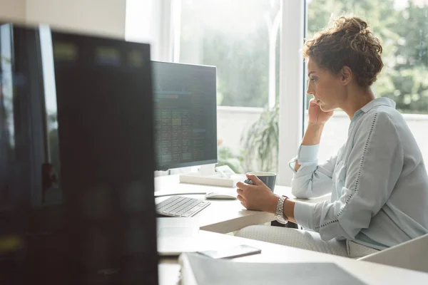 年轻的女实业家在现代办公场所的台式电脑上工作时喝咖啡 — 图库照片