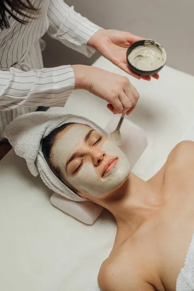 白人美容师妇女在一个美丽的年轻白种女人的脸上涂抹面部面具躺在按摩床上 — 图库照片