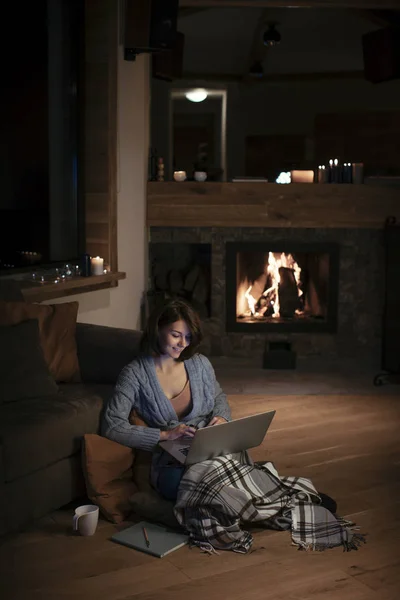 美丽的微笑的女人坐在木地板上的壁炉和打字她的笔记本电脑 — 图库照片