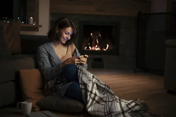 美丽的微笑的女人坐在壁炉和打字的智能手机 — 图库照片