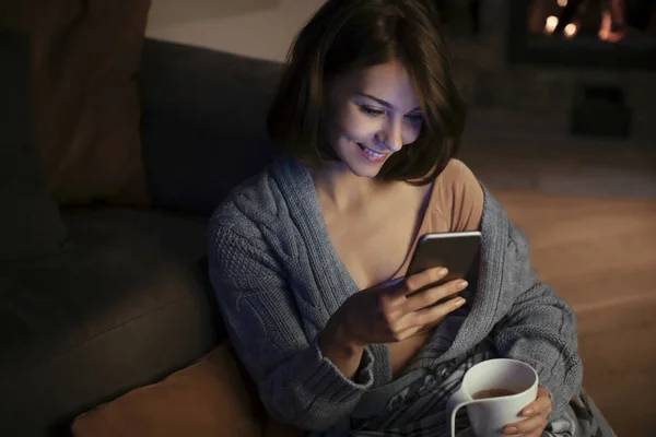 美丽的微笑的女人坐在壁炉和打字的智能手机 — 图库照片