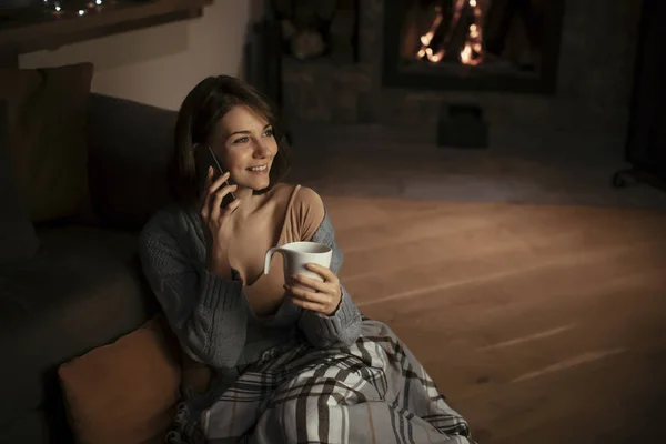 Beautiful smiling woman sitting by the fireplace and talking on smartphone.