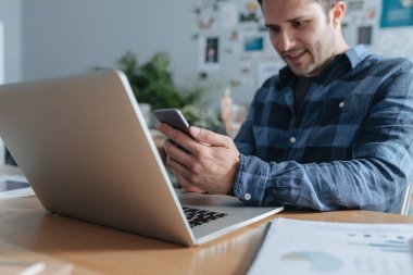 Yakışıklı genç gülümseyen beyaz adam çalışma masasında cep telefonunu kullanma.