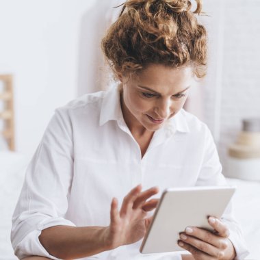 Yatağında oturmuş ve tablet kullanarak güzel gülümseyen beyaz kadın.