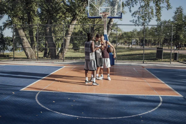 Group of young  basketball players standing on outdoor court and looking at smartphone.