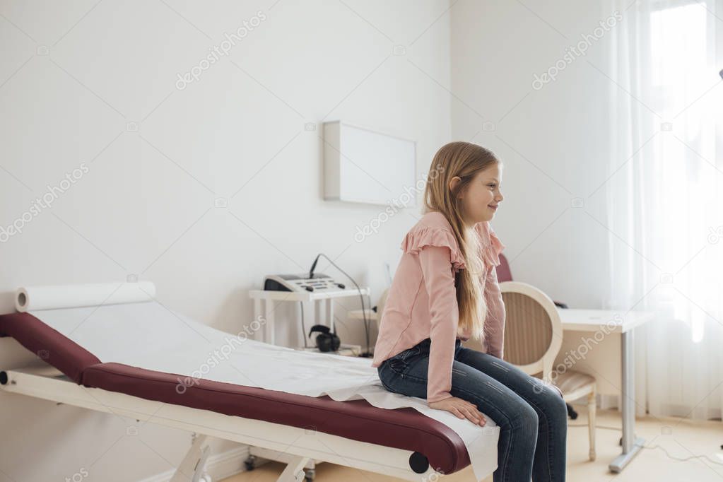 Cute Caucasian girl sitting at examination room at the clinic and waiting for a doctor.