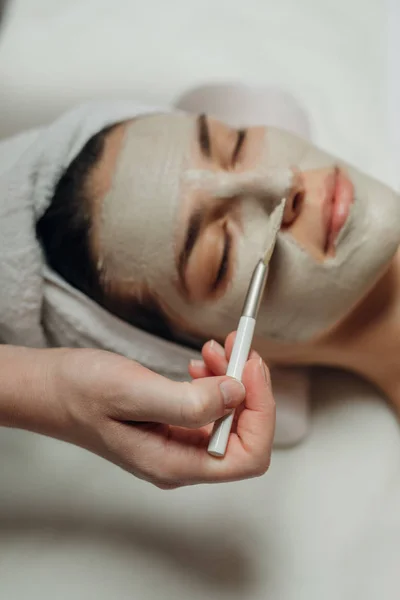 マッサージ ベッドに横になっている美しい若い白人女性の顔に顔のマスクを適用する白人美容師女性の手 — ストック写真