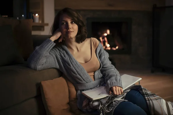 かなり白人女性は暖炉のそばで居心地の良いセーターに座ってノートを保持しています — ストック写真