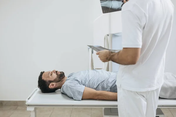 Jovem Médico Examinando Filmes Raios Enquanto Seu Paciente Está Deitado — Fotografia de Stock