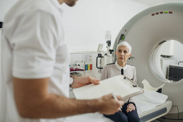 Doctor talking to senior patient and taking notes next to a CT scan machine.