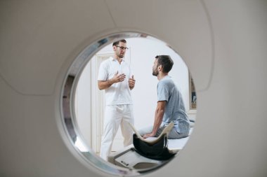 Young medical technician talking to his patient sitting on the CT scanner bed. clipart