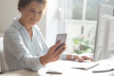 Genç işkadını modern ofis alanı, bir masaüstü bilgisayarda çalışırken bir cep telefonu kullanarak.