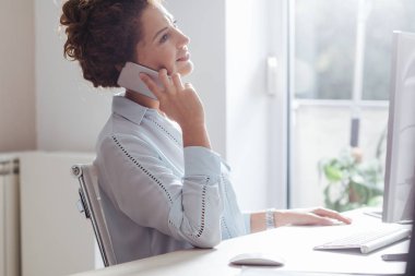 Genç işkadını modern ofis alanı, bir masaüstü bilgisayarda çalışırken bir cep telefonu kullanarak.