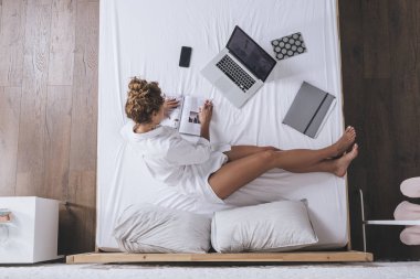 Kadın fotoğrafı yukarıda bir kitap okuma ve onun yatağında okurken onu laptop yazma.