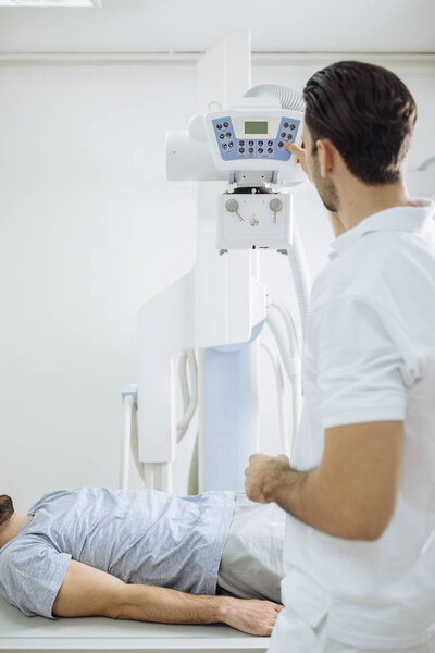 Young medical technician operating an X-ray machine while his patient is lying on the bed in front of him in a clinic.