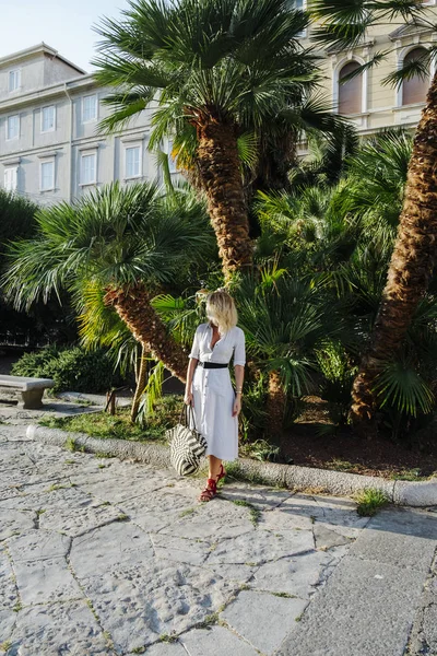 Güzel ve zarif şık beyaz bir kadın çantasını palmiye ağaçları önünde poz Ise tutarak güneş gözlüğü ile.